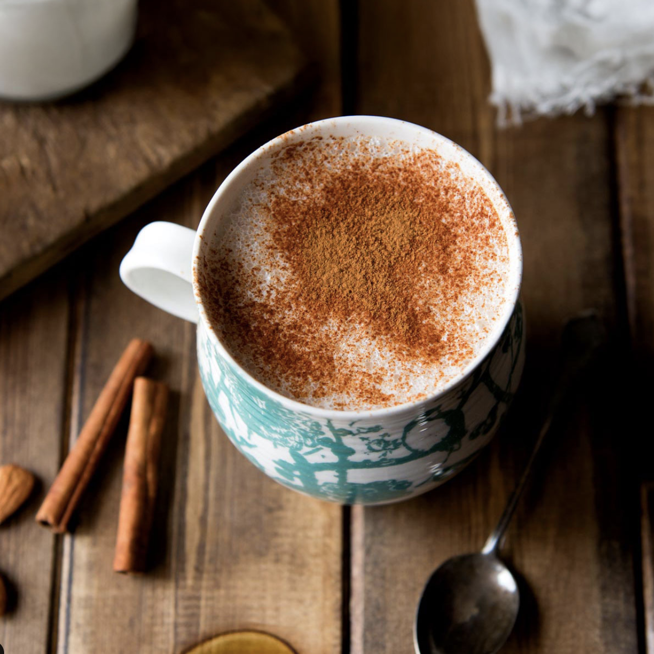 حليب دافئ و رشة قرفة عالوجه في كوب ابيض مزخرف بالازرق على طاولة خشبية