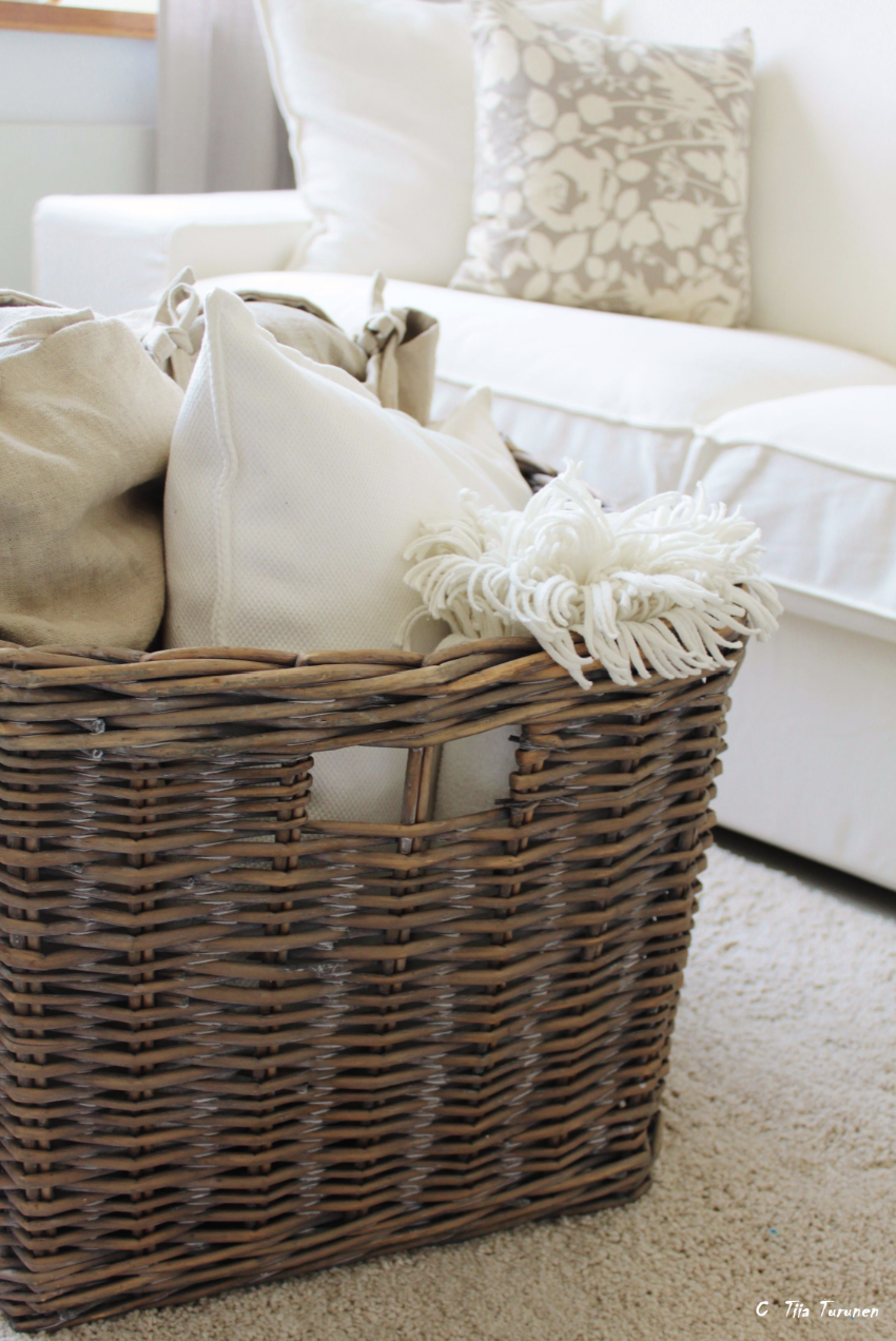 سلة خوص كبيرة بجانب كنبة فاتحة تحتوي بطانيات ووسائد، تعطي لمسة عضوية دافئة في غرفة معيشة حيادية.