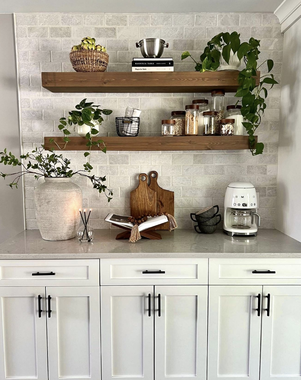 رفوف خشبية مفتوحة فوق كاونتر قهوة مع سلة خوص ونباتات خضراء ولمسات داكنة بسيطة، بتنسيق مرتب بدون زحمة