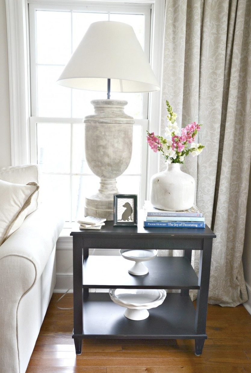 طاولة جانبية صغيرة عليها مصباح طويل، كتابان مكدسان، ومزهرية بيضاء متوسطة تضيف ارتفاعًا وتناسقًا.