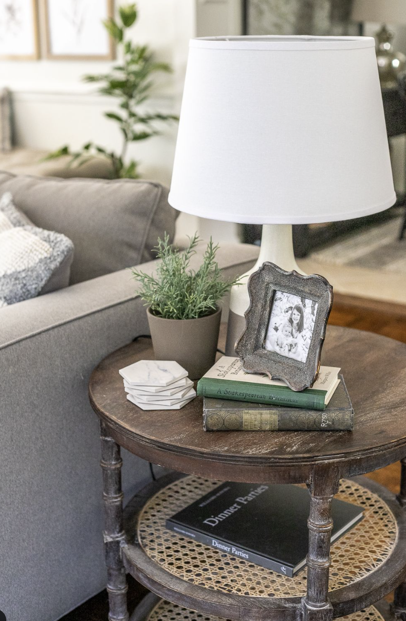 طاولة جانبية دائرية بجانب كنبة عليها مصباح أبيض، كتابان مكدسان، ونبتة صغيرة تضيف توازنًا بصريًا.