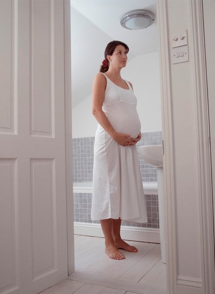 حامل تقف في حمام بإضاءة طبيعية هادئة استعدادًا لدش دافئ يساعد على الاسترخاء أثناء الحمل.